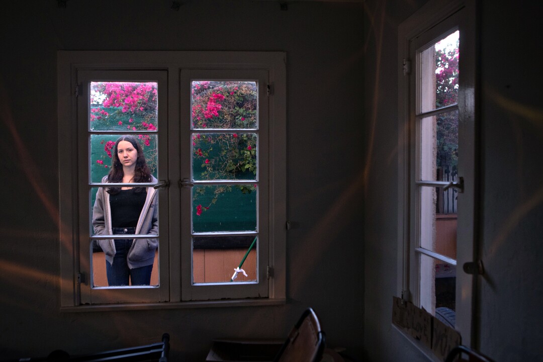 A young woman stands on the patio and looks into a home through a window. 