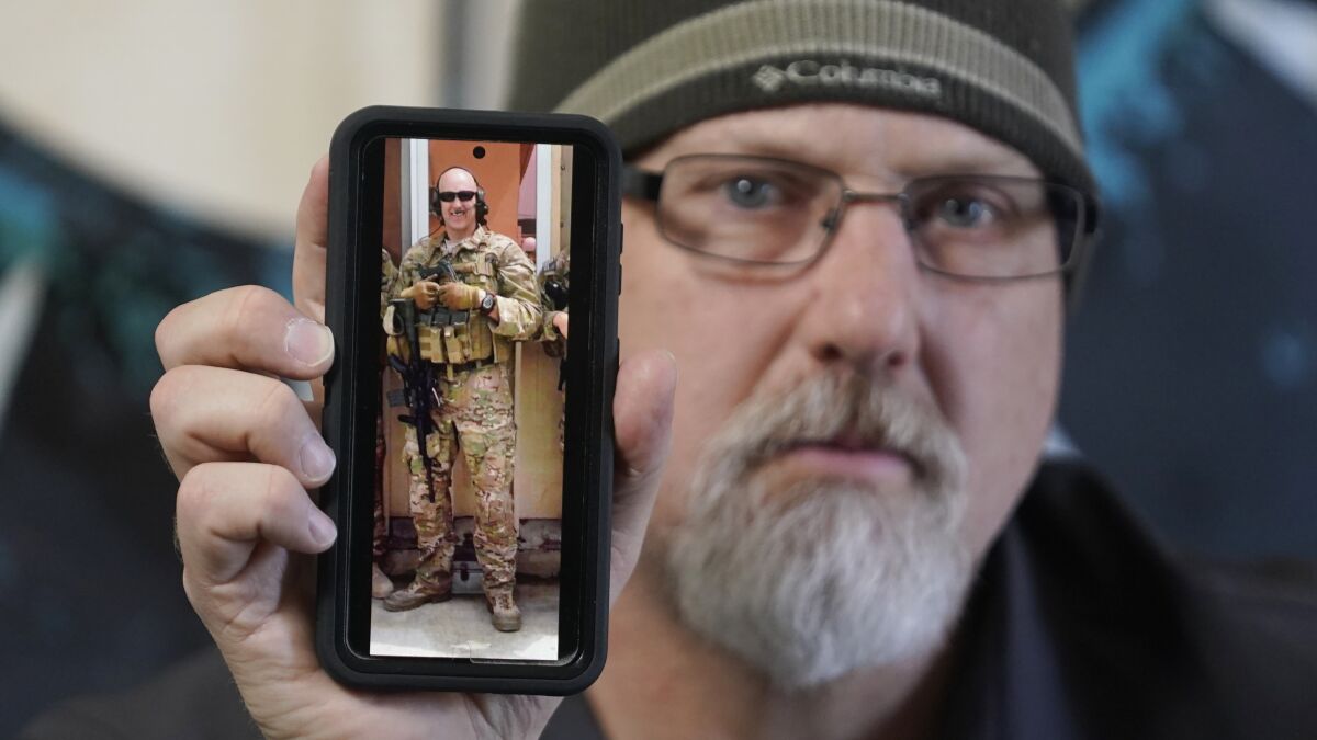 A bearded man holds a phone with a photo on the screen of himself in military gear