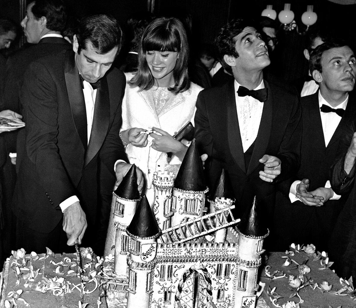 A man, standing next to a woman and two other men, cutting a large cake in the form of a castle