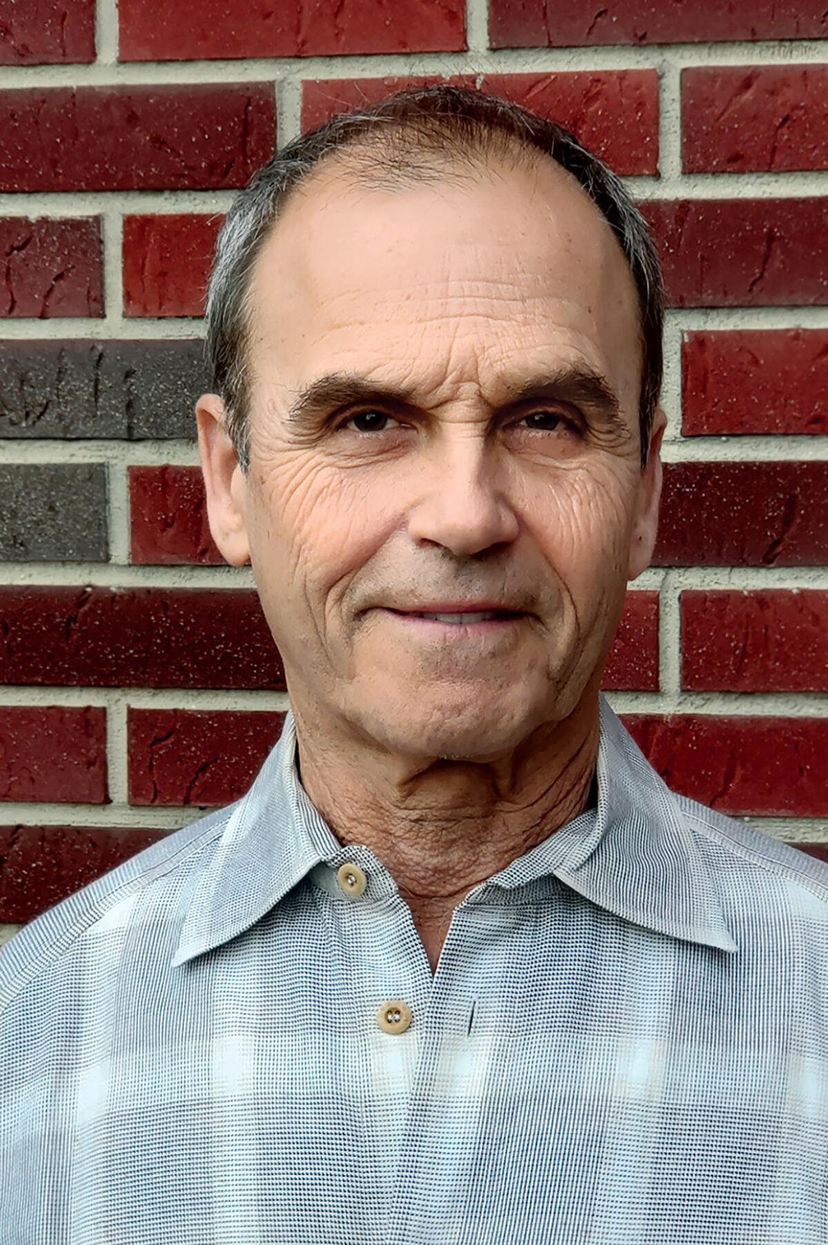 A balding man in a windowpane-checked shirt stands in front of a brick wall.