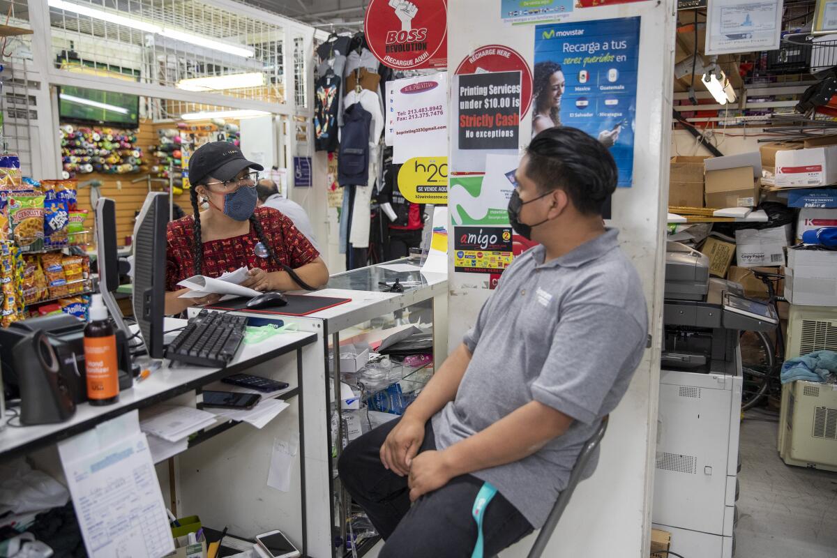 Two people wearing masks talk across a counter in a cluttered store.