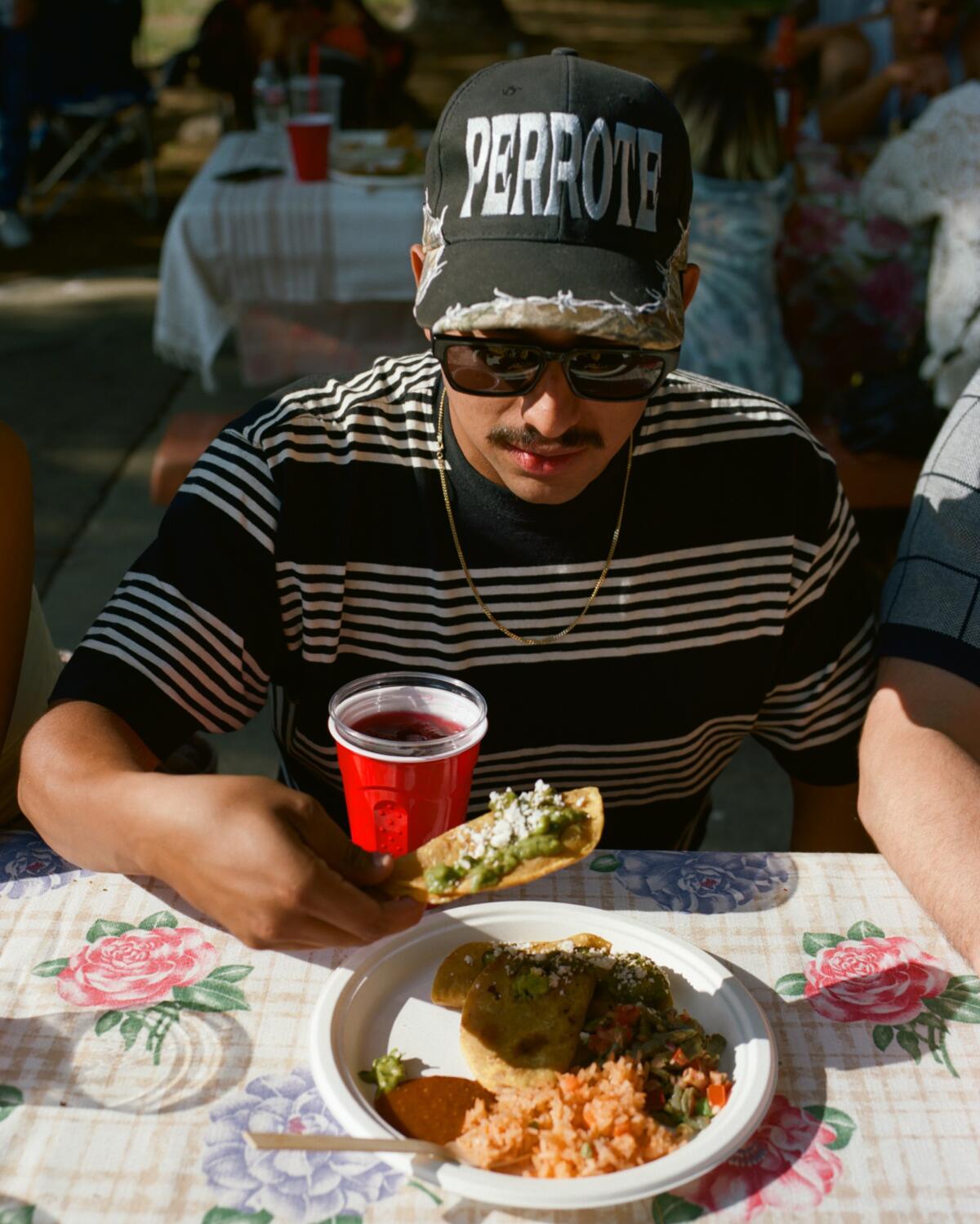 a man wearing sunglasses and a hat begins to eat a taco