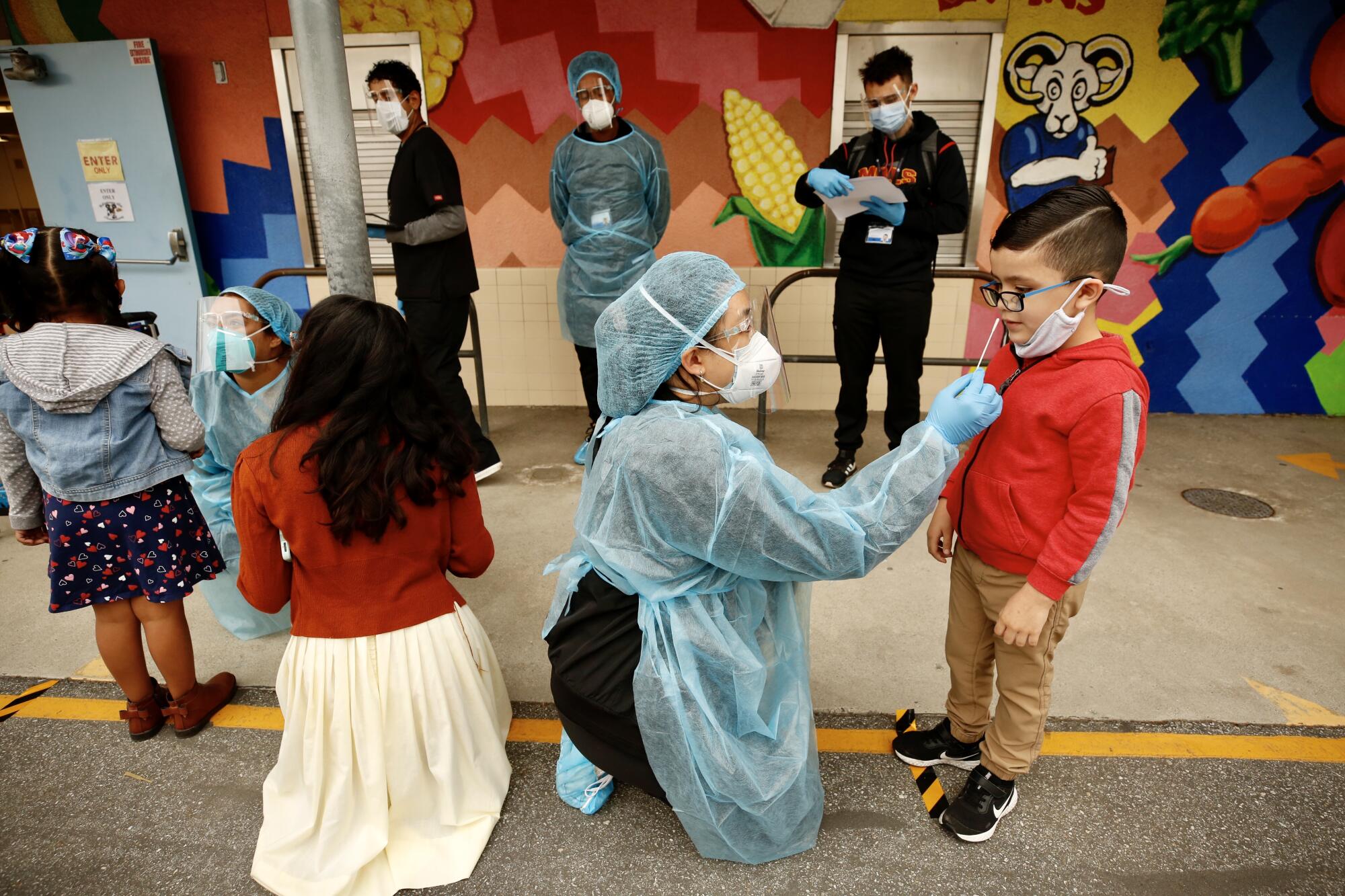Women in protective gear kneel in front of young students to conduct nasal swab tests.