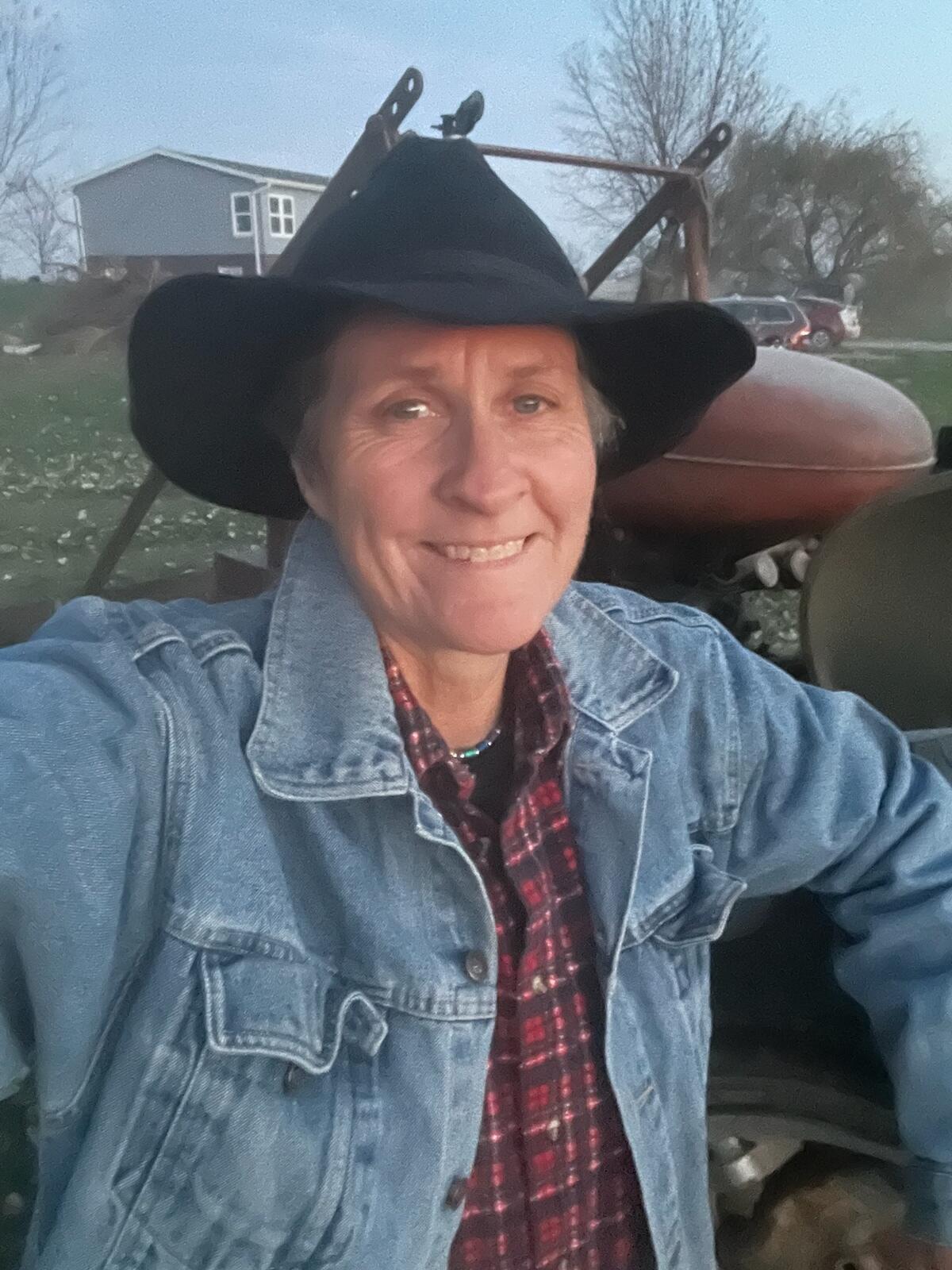 A woman in a red plaid shirt, denim jacket and black hat, pictured with farm equipment on land with a house in the distance