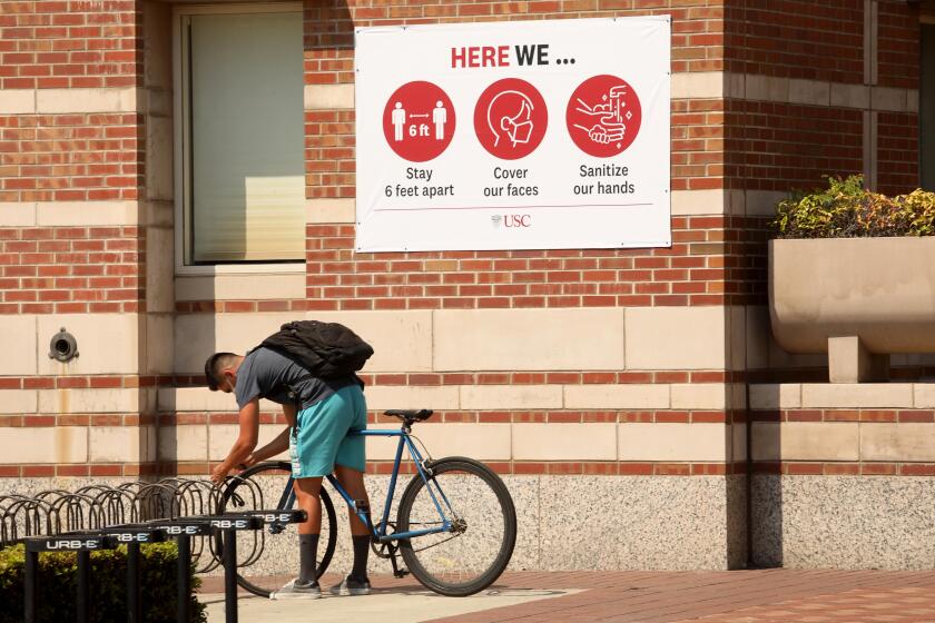 LOS ANGELES, CA - AUGUST 17, 2020 - - USC student Jesus Carreon, 21, locks up his bike under a sign that states three steps to keep the coronavirus at bay on the first day of academic instruction for the Fall 2020 semester on the USC campus in Los Angeles on August 17, 2020. Carreon wanted to do his homework on a nearby patio to have some semblance of a college experience. Students were taking online courses off campus due to the coronavirus pandemic and the university was nearly empty. (Genaro Molina / Los Angeles Times)