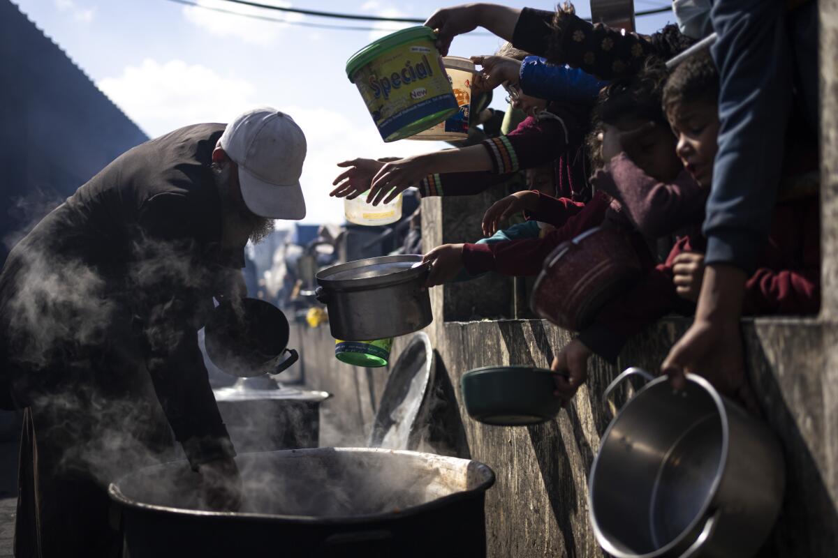 People hold out pans as a person scoops out hot food from a huge container in Rafah, Gaza Strip.