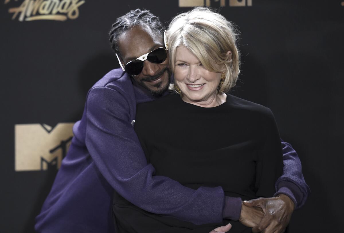 Snoop Dogg, clad in sunglasses and a purple top, embraces Martha Stewart from behind and presses his cheek to hers