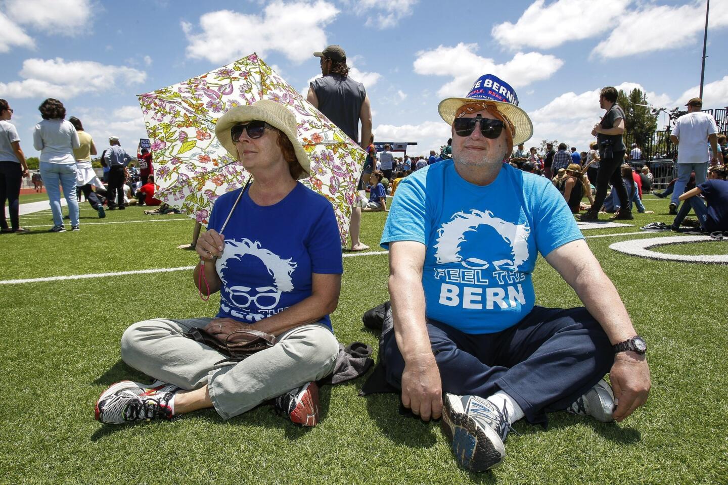 Robin Hampton and Jim Drogo, both from Fallbrook, sit on artificial turf as they and others wait to see Democratic presidential candidate Bernie Sanders.