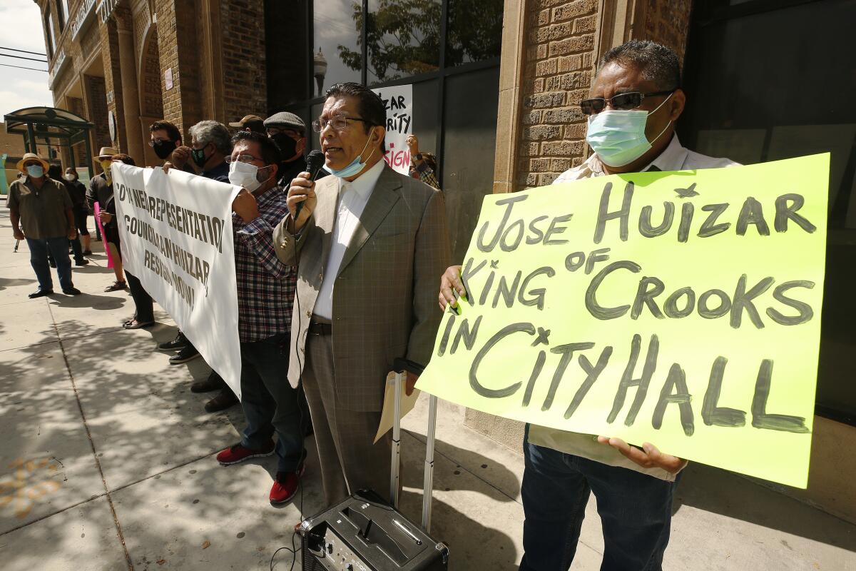 Men hold signs outside a building, one in focus reading "Jose Huizar King of Crooks in City Hall."