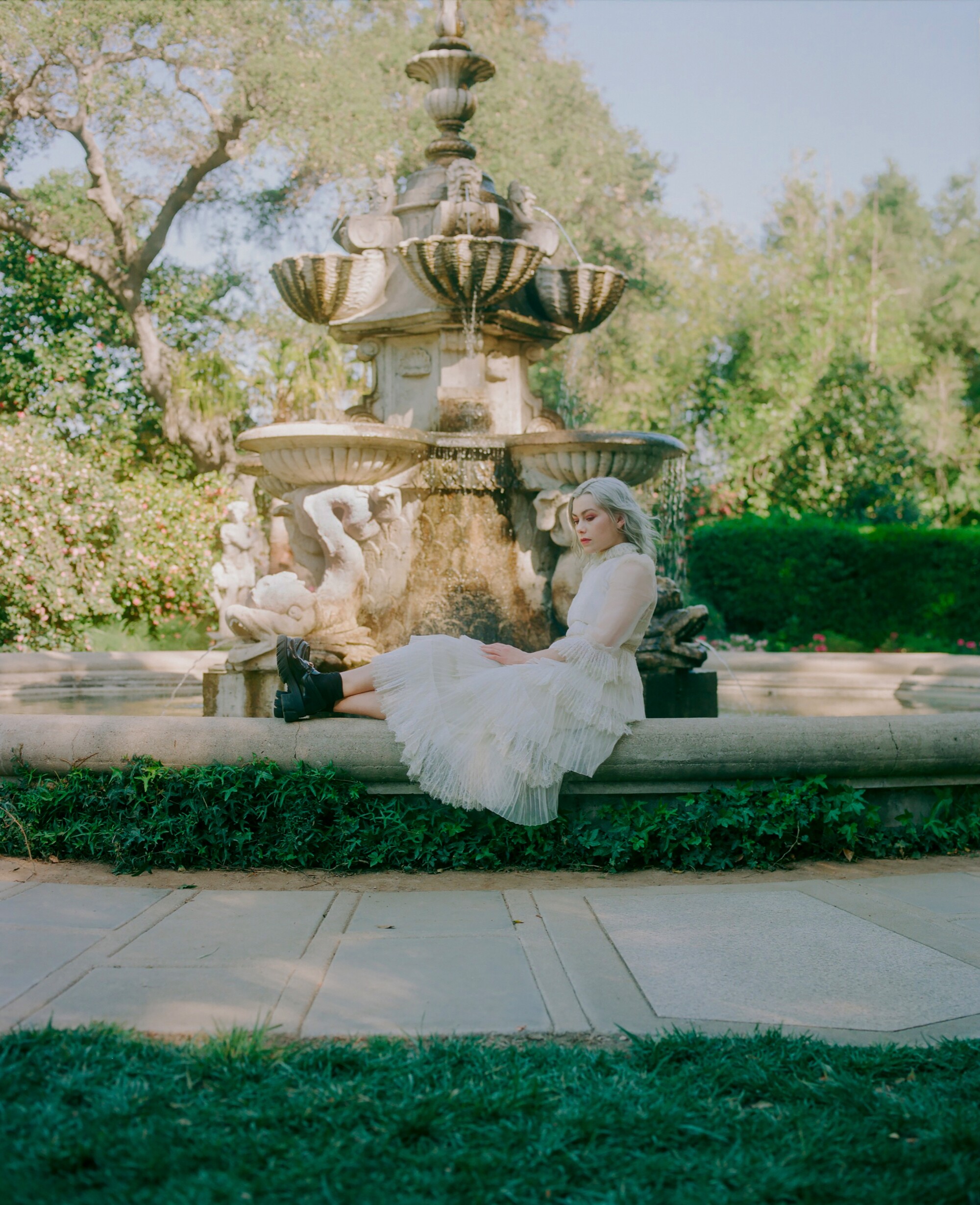 A woman in a white dress sits near a grand outdoor fountain.