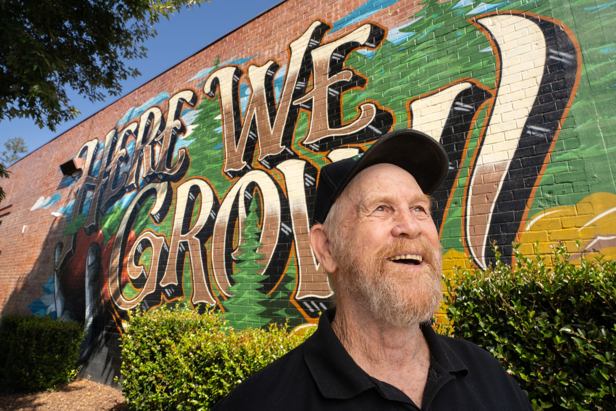 A bearded man smiles in front of a mural.
