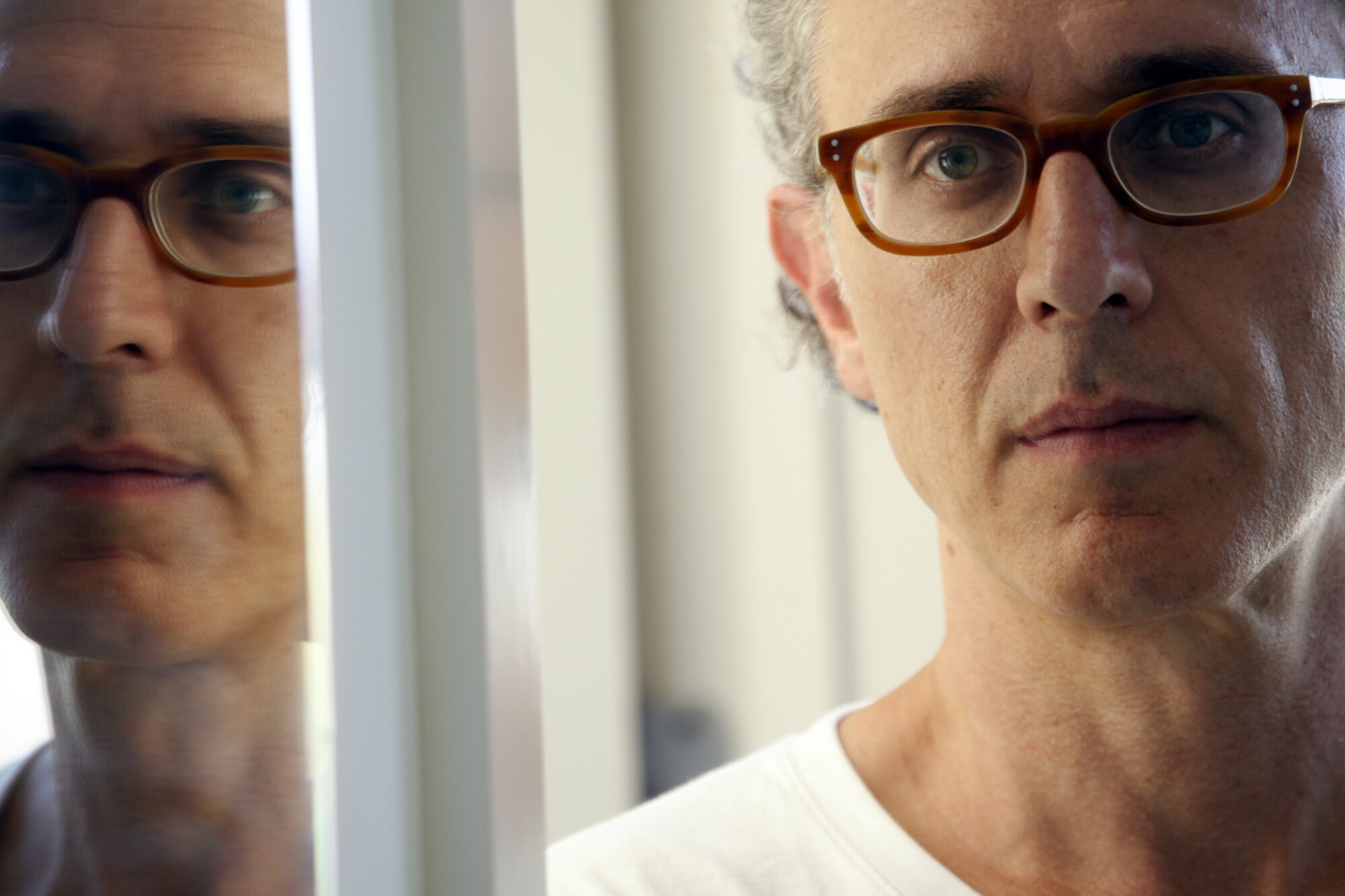 A man with orange-rimmed glasses and a white T-shirt stands next to a mirror