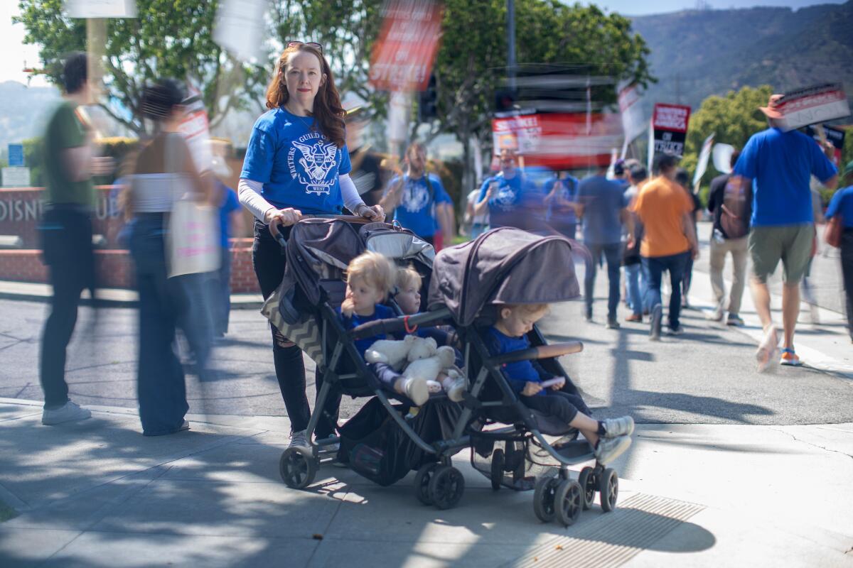 A woman stands behind a stroller seating three young children as striking writers walk behind her.
