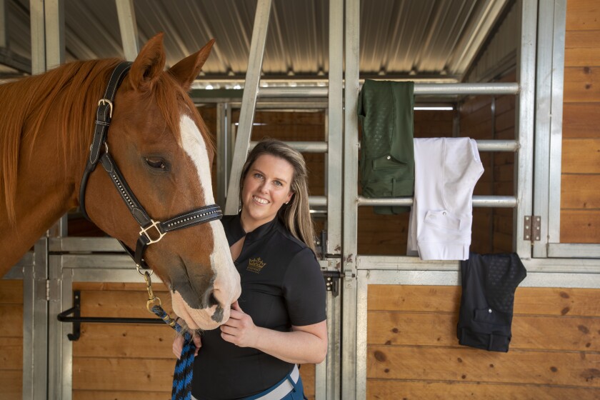 A woman stands near a horse while horse-riding pants hang near her.