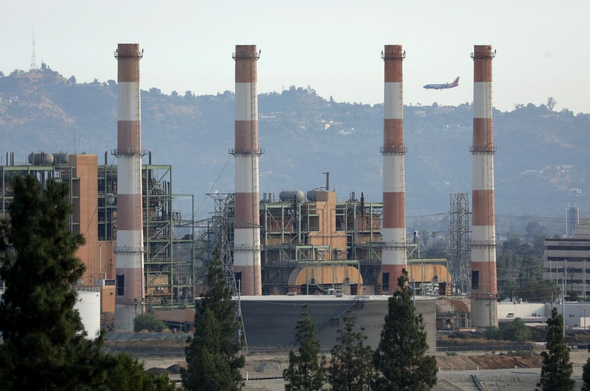 A plant's four smokestacks are seen. In the back are hills, homes and hazy air.