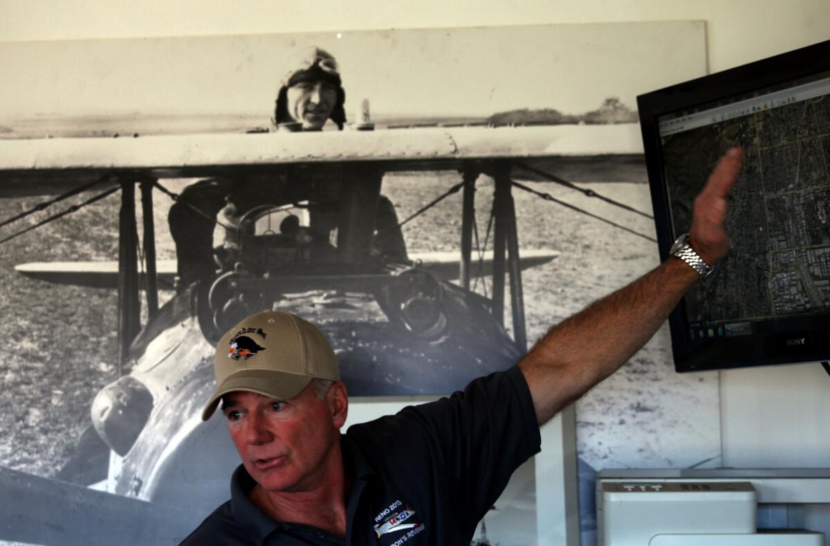 A man in a billed cap stands in front of a vintage photo of a pilot in a plane as he gestures toward a screen.