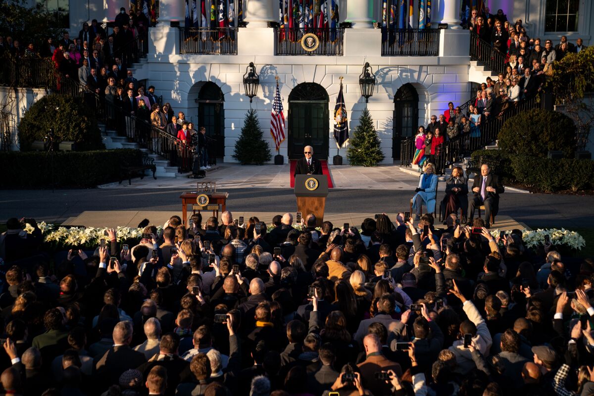 President Joe Biden stands at a microphone in front of the white house, surrounded by people and rainbow lights