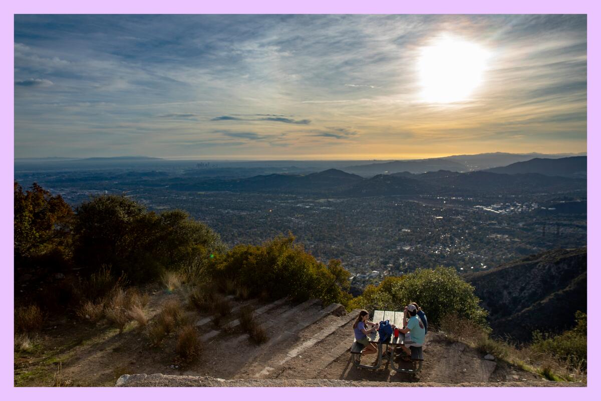 An aerial view of three hikers sitting at a picnic table atop a mountain with the sun setting in the distance