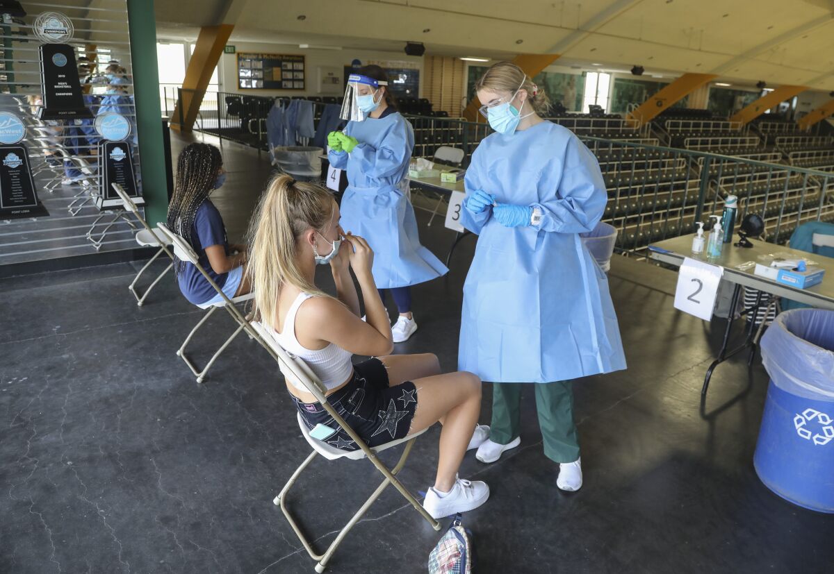 Workers in blue gowns and masks prepare to give coronavirus swab tests to two women in a college gymnasium