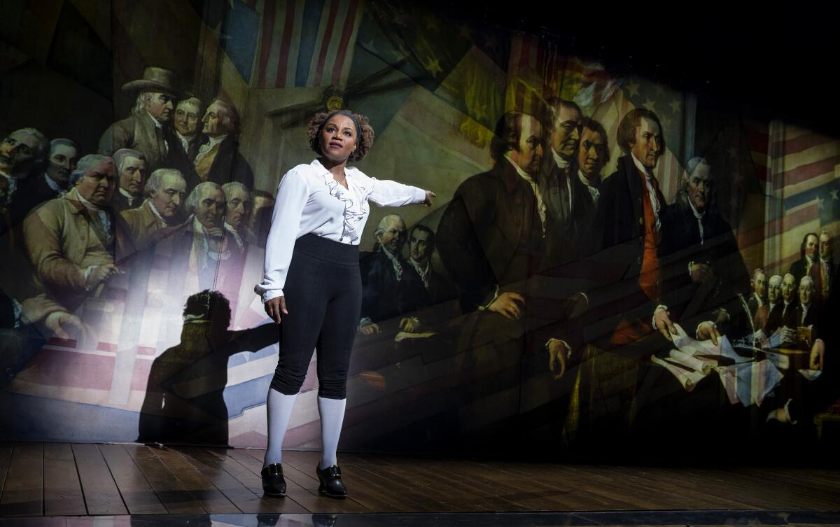 Gisela Adisa, dressed in period attire, points to a projected painting of the Founding Fathers.