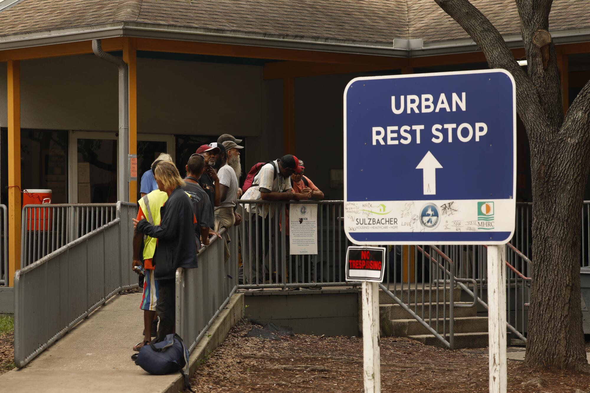 People stand near railings on a walkway to a building, near a blue-and-white sign that reads Urban Rest Stop