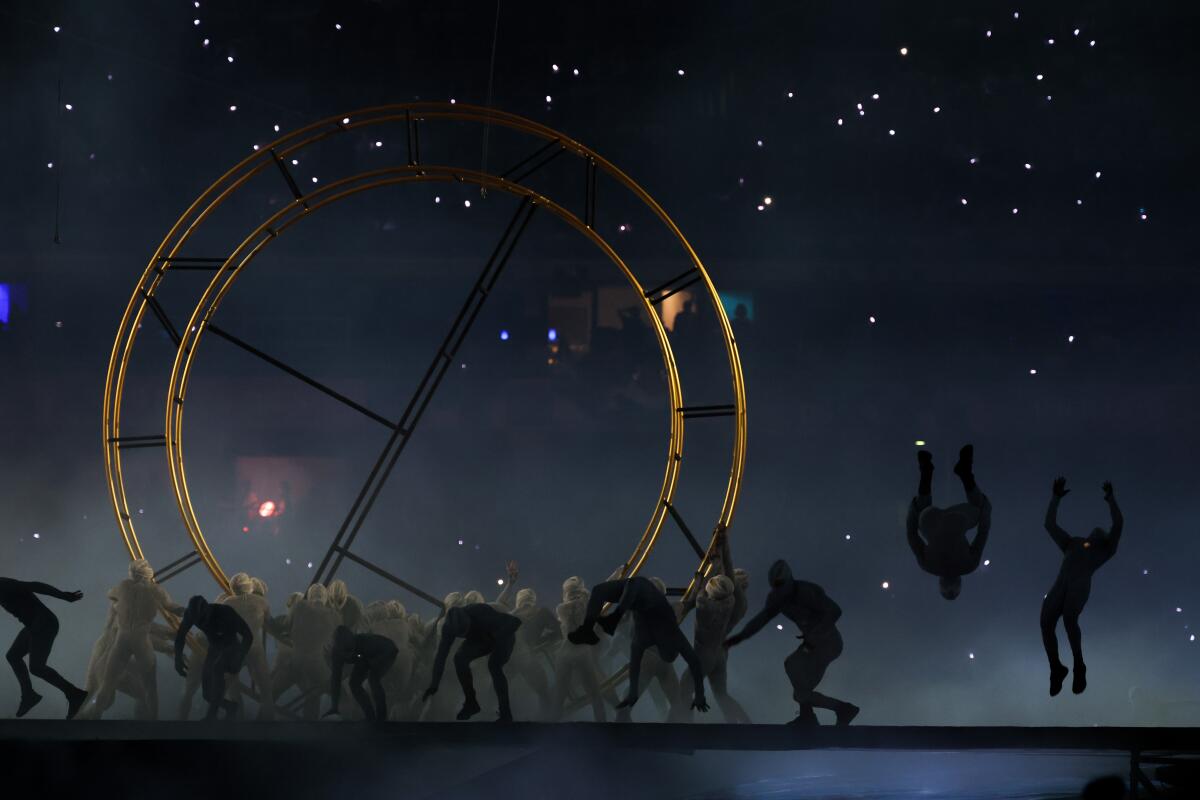 People in silhouette moving in front of a giant, gold-colored ring.