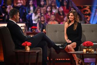 Jesse Palmer, left, wears a black suit and Leslie Fhima, right, wears a black dress as the two sit on grey couches