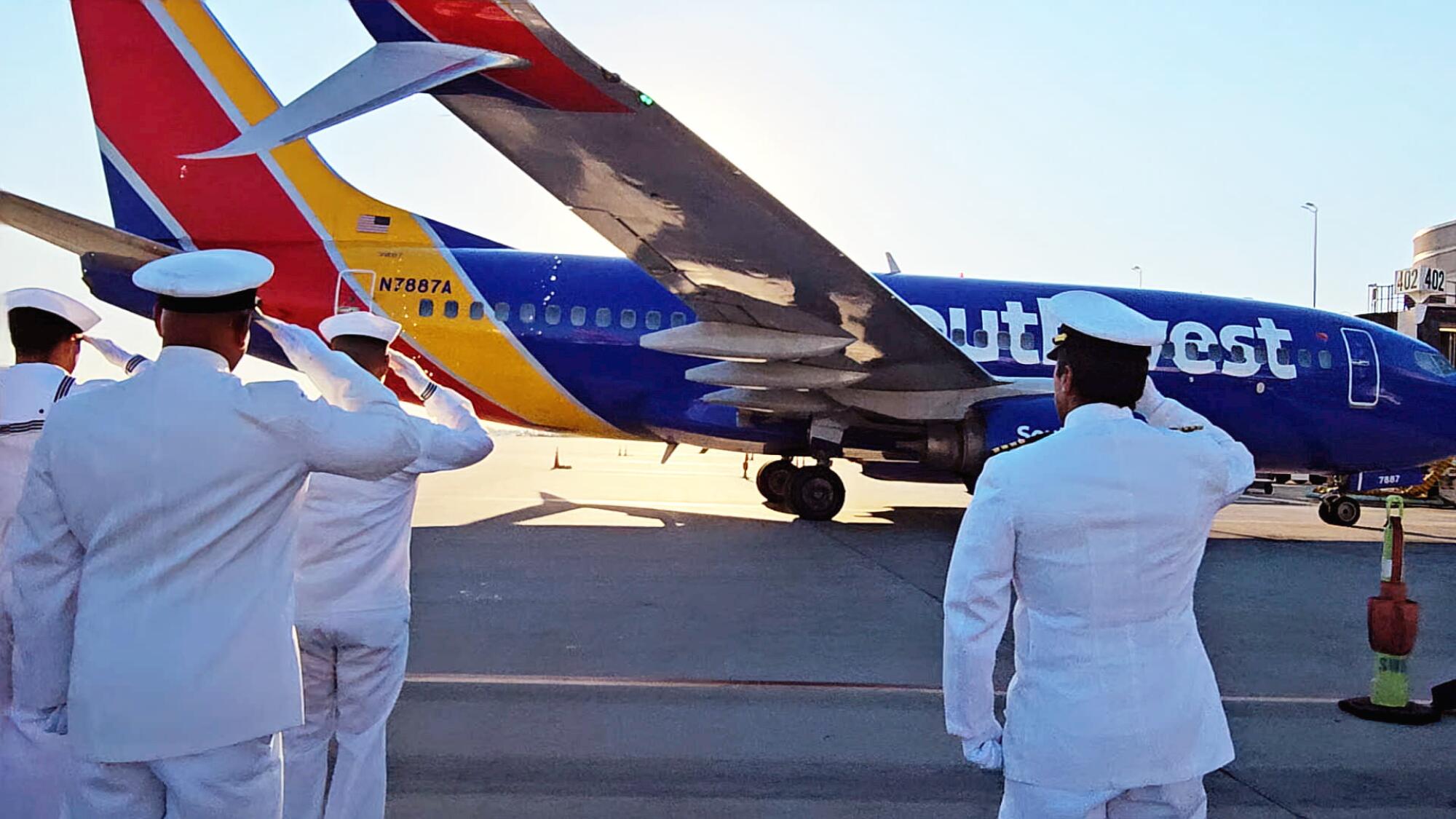 Men in white hats and uniform salute while standing before a red, yellow and blue aircraft on the tarmac