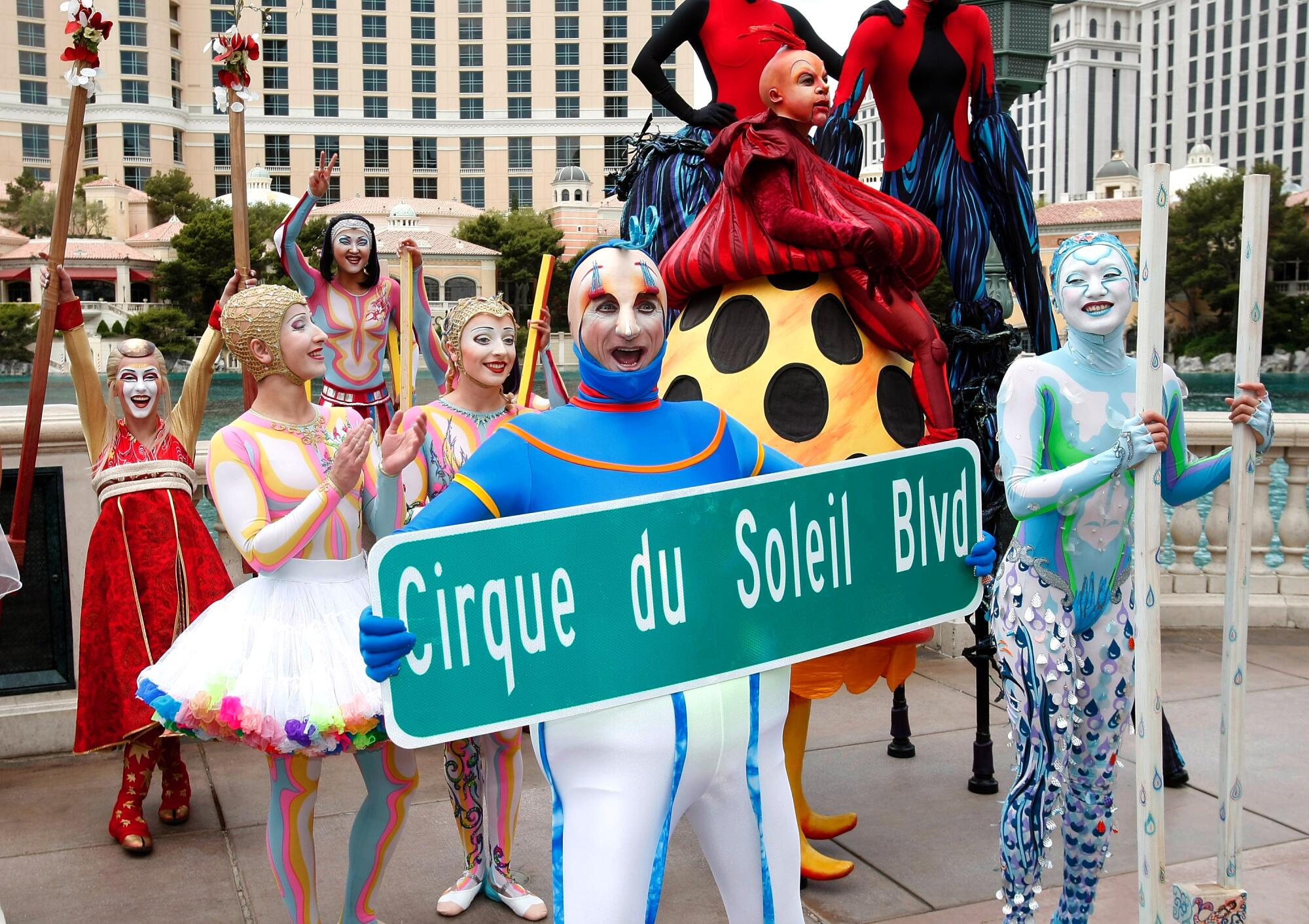 A performer holds a street sign with the words "Cirque du Soleil Blvd" as other performers stand nearby.