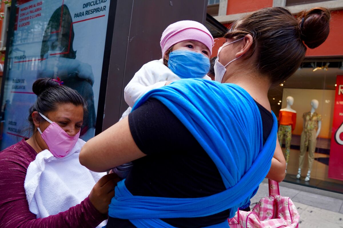 A woman holds a baby in a pink cap while another woman, left, examines the child. All three are wearing masks