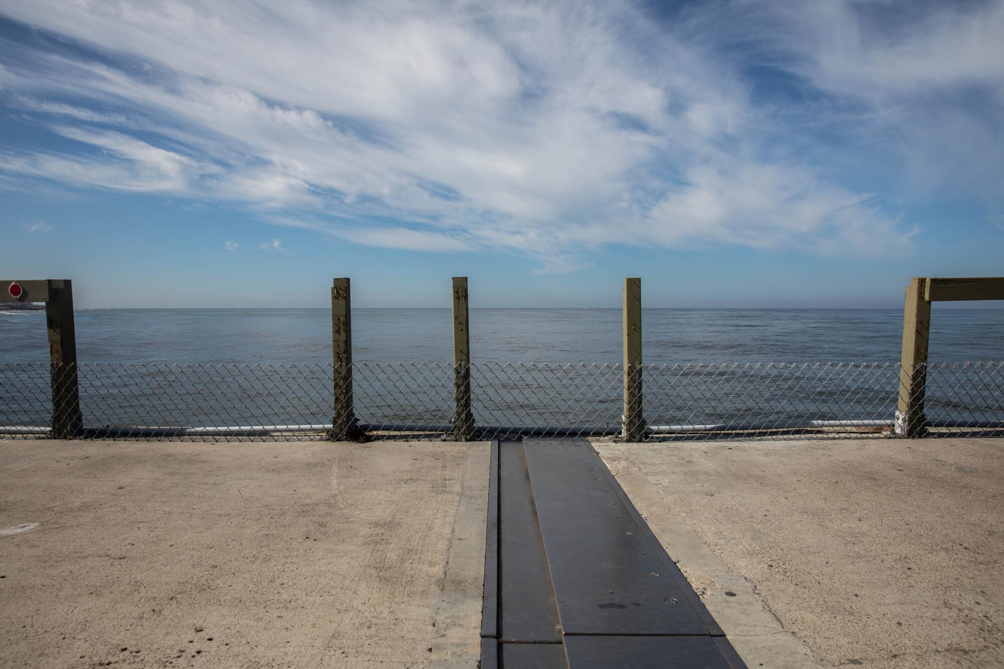 A calm ocean and cloud-streaked blue sky are seen beyond the edge of a pier, where railings are missing from posts.