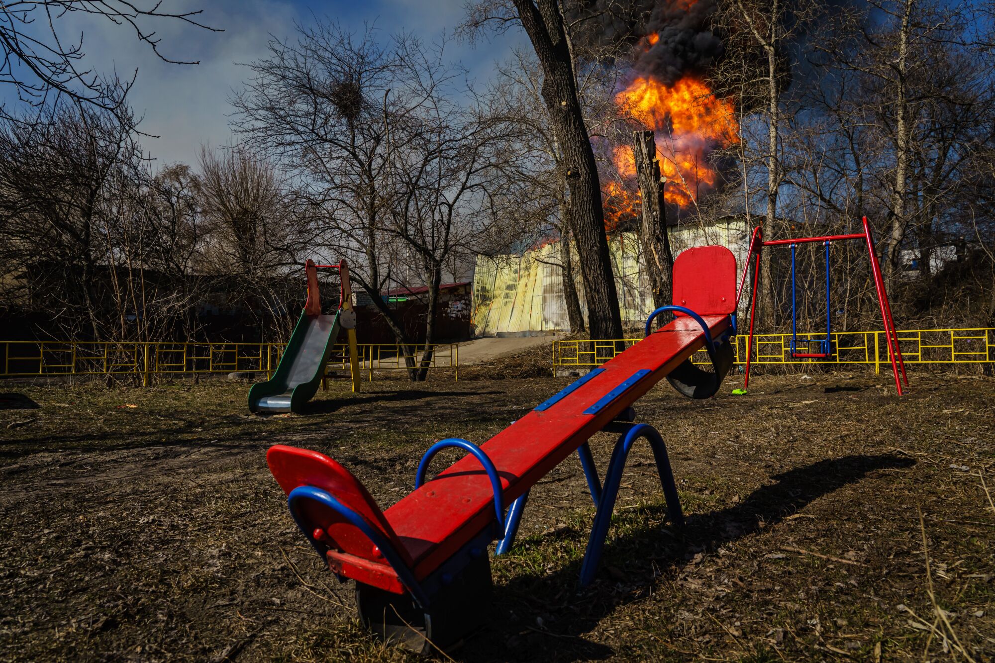 A red seesaw on a playground in the foreground with a building on fire in the background.