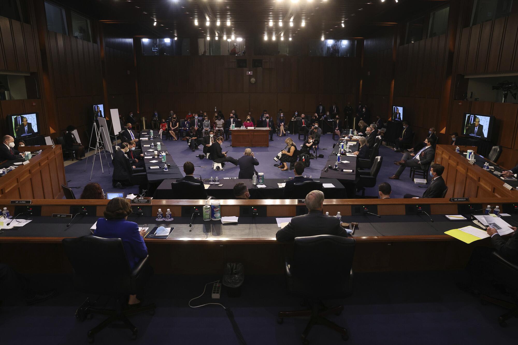 An overview of the hearing room shows senators, spaced a few feet apart, and Amy Coney Barrett seated at a desk.