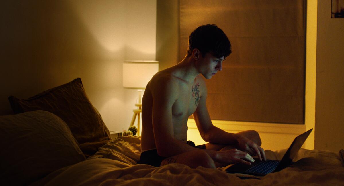 A bare-chested man types on his laptop in bed in a darkened room.