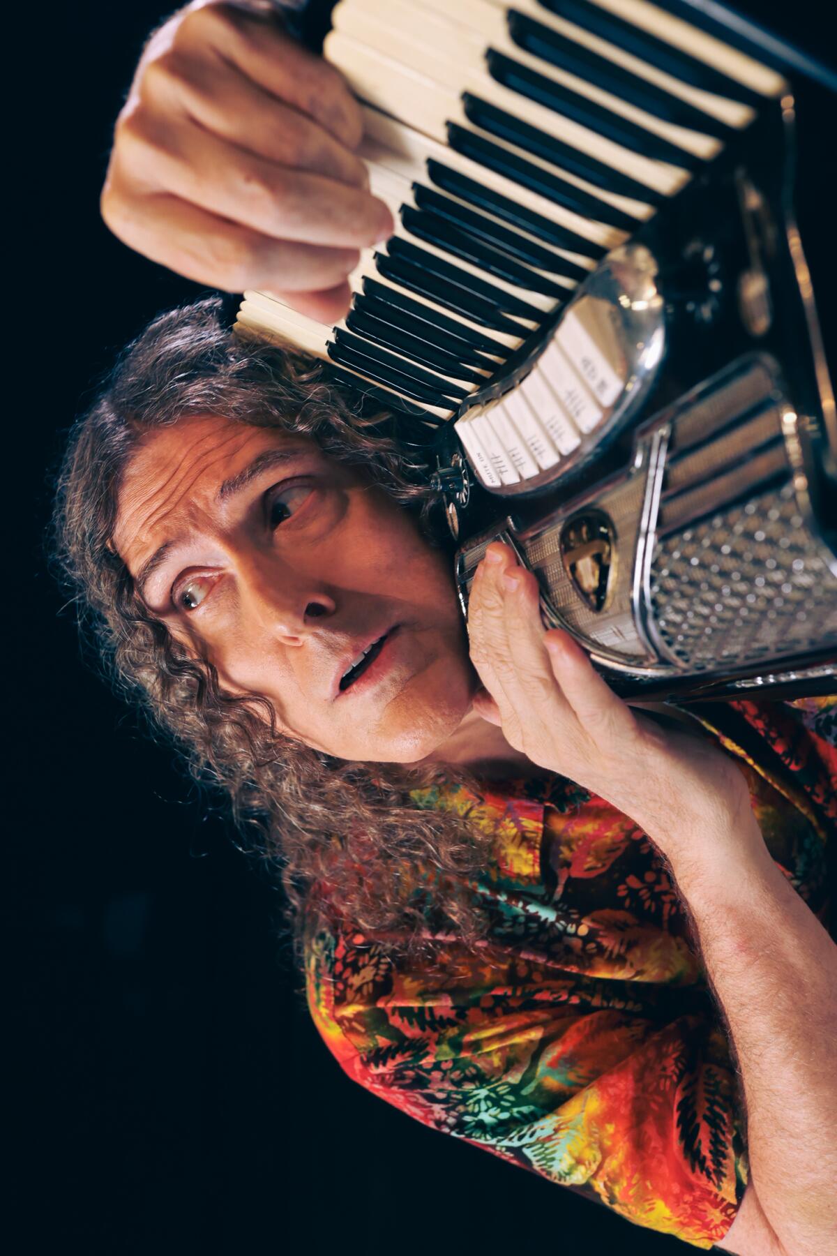 A long-haired, colorfully dressed man lovingly embracing his accordion, staring emotively off in space