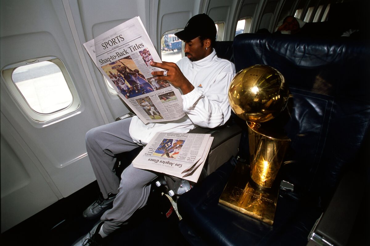 Kobe Bryant sits on an airplane next to a trophy, holding the L.A. Times Sports section.