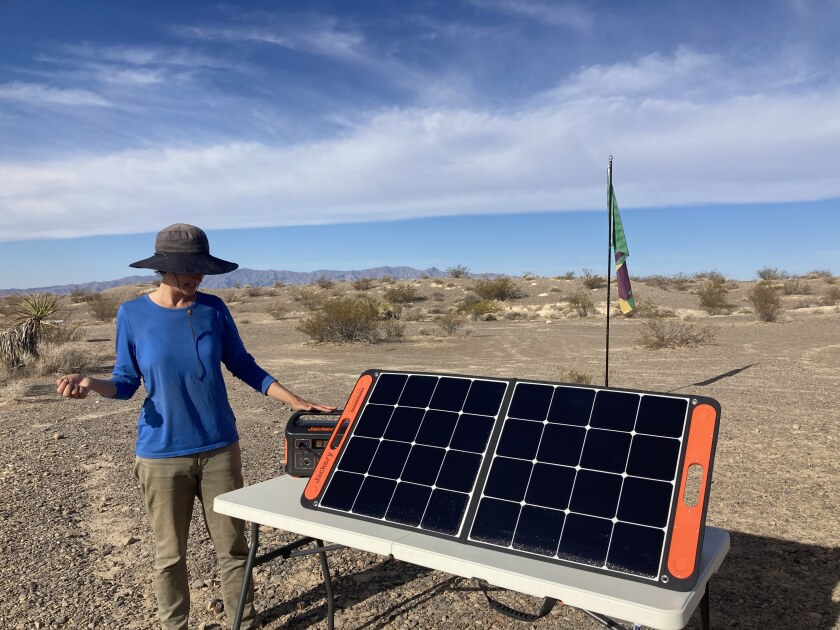 A woman in a hat and blue top and khaki pants stands next to a table with a solar panel