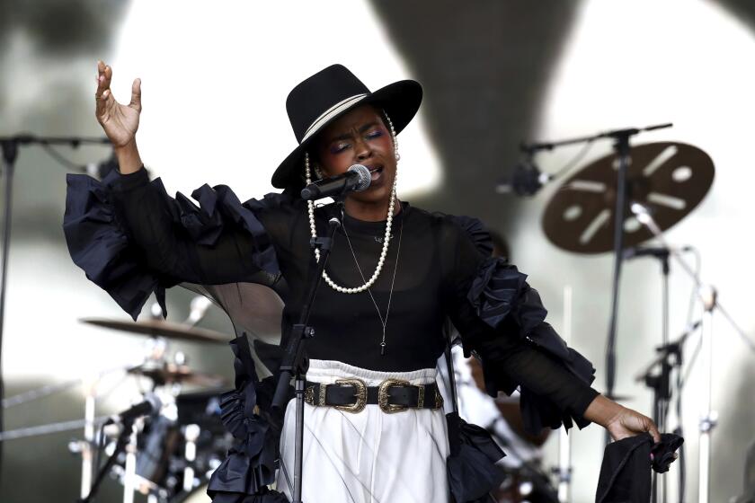 A woman in a black hat, black shirt and white pants singing in front of a microphone on a stage