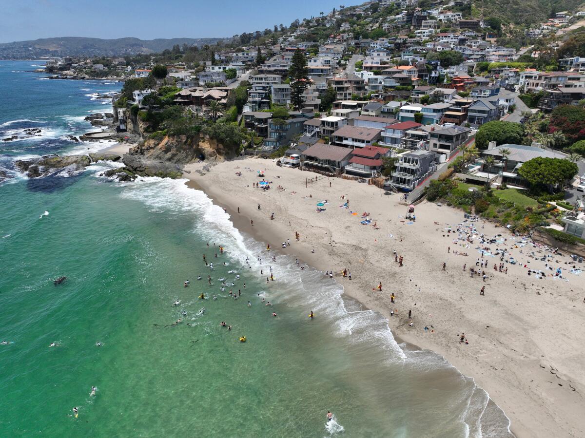 An aerial view of Victoria Beach, with swimmers in the green water and sunbathers on the sand against a residential hillside.