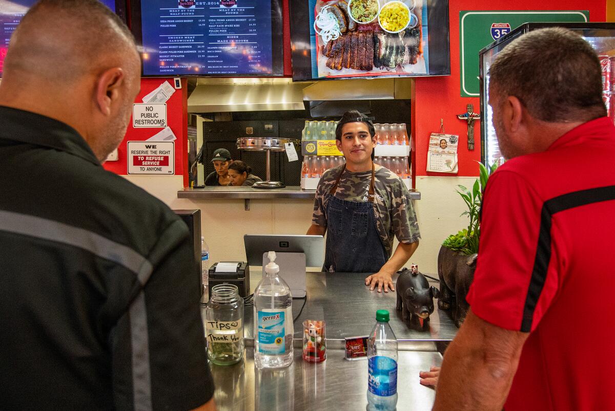 A man behind a restaurant counter talks to two men, their backs to the camera