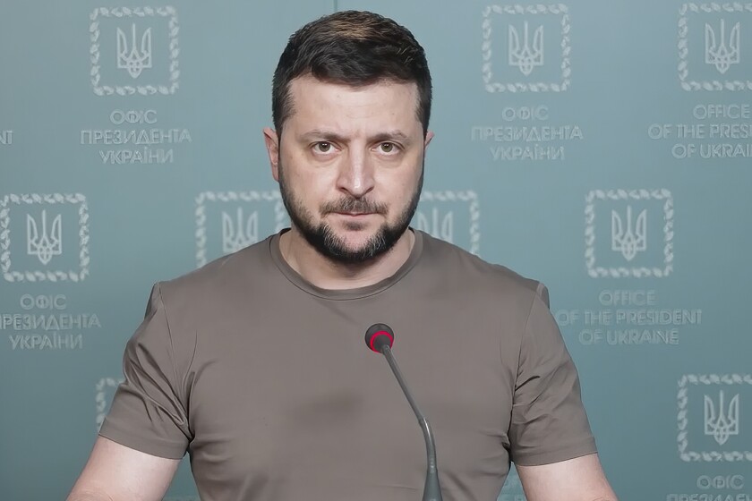 A man with dark hair and a beard, wearing a brown T-shirt, speaks before a microphone