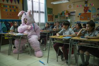 Renaldo wears a large pink bunny suit with a unicorn horn while seated amid children in a classroom in "Los Espookys."
