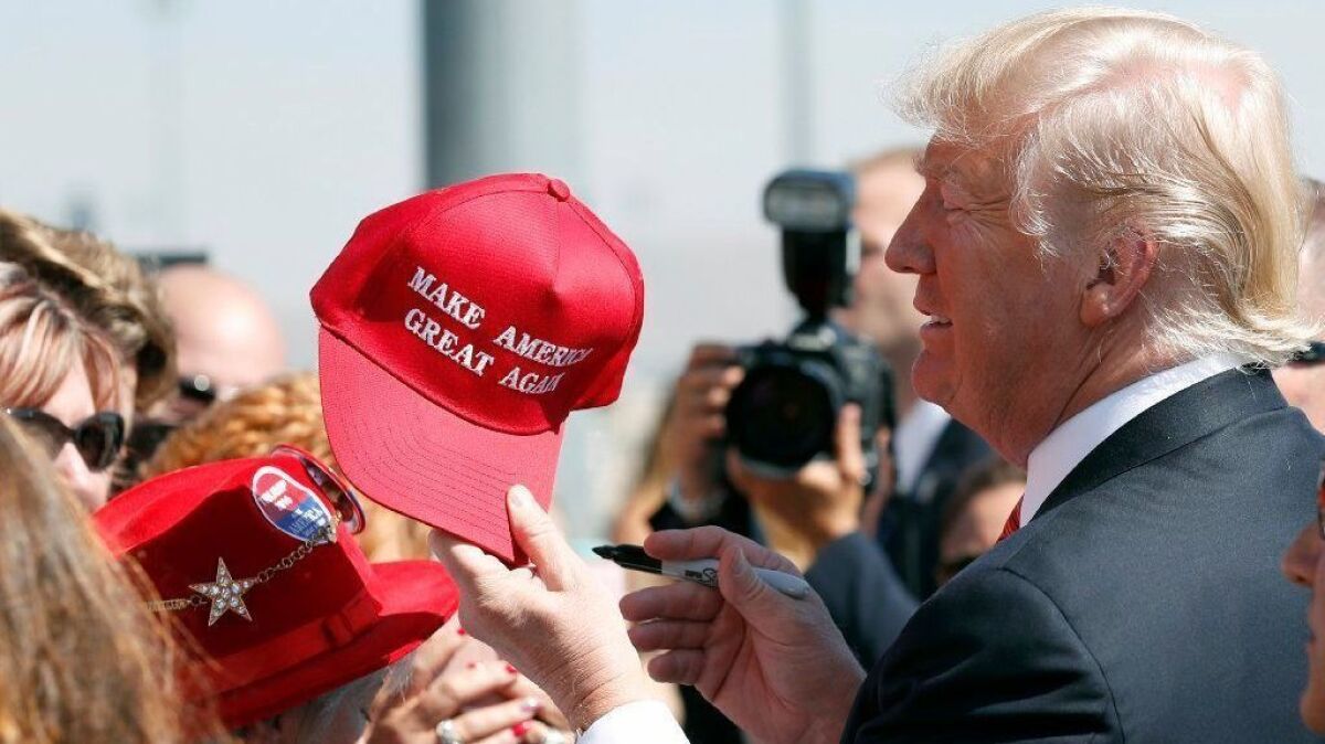 Donald Trump stands among supporters holding a red ball cap with the words "Make America Great Again" on it.