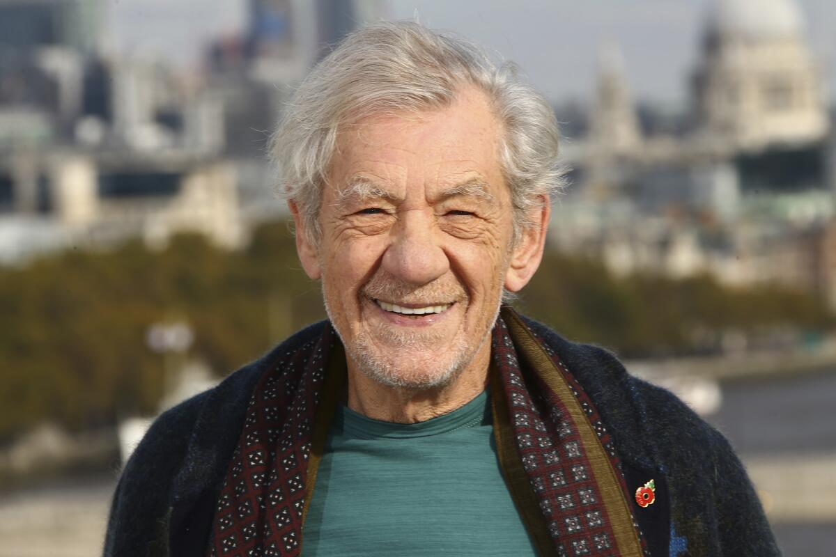  Ian McKellen wearing a blue-green T-shirt and a printed scarf with a dark cardigan outdoors in front of a cityscape
