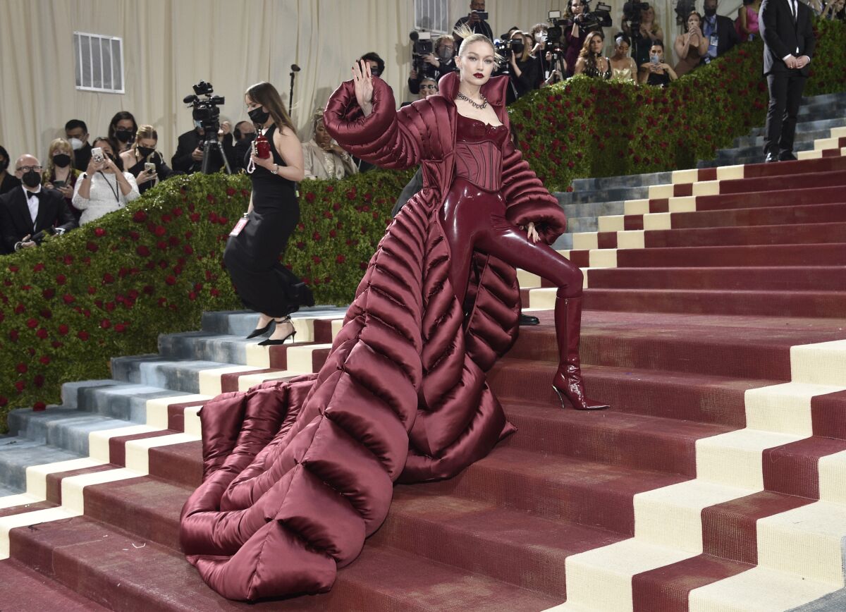 A woman in cranberry leather pants and a bustier plus a massive long coat stands on stairs at an event