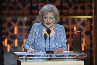 An older woman with white hair in a blue outfit speaks at an awards show lectern
