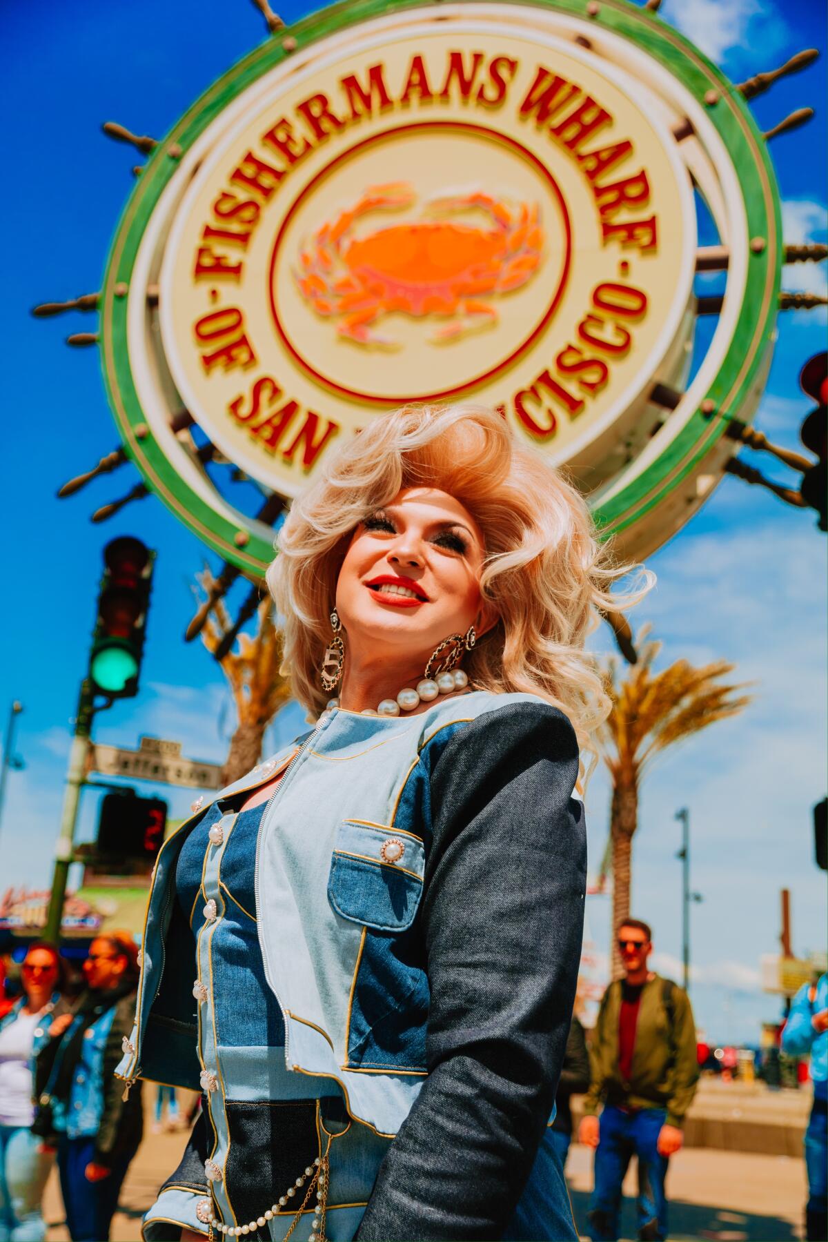 A person with blonde hair poses for. photo in front of a sign that reads "Fisherman's Wharf Of San Francisco."