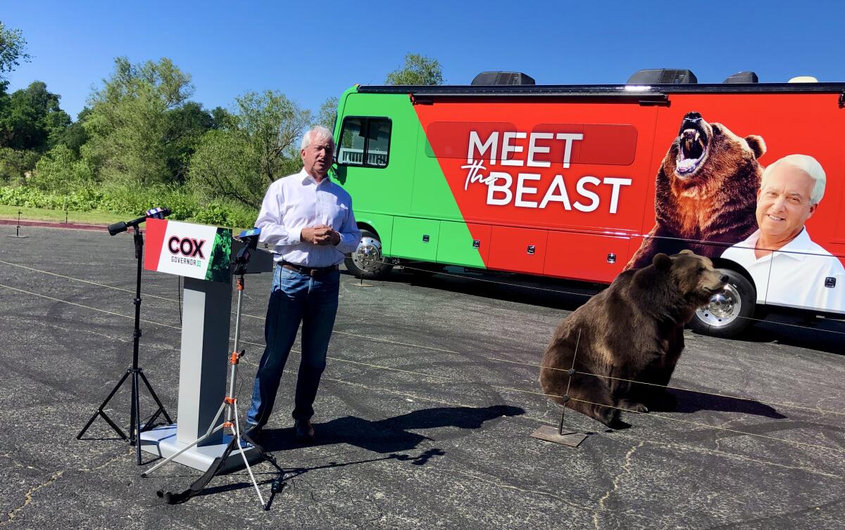 John Cox stands behind a podium with a bear on his right and a bus behind him that says "Meet the beast"