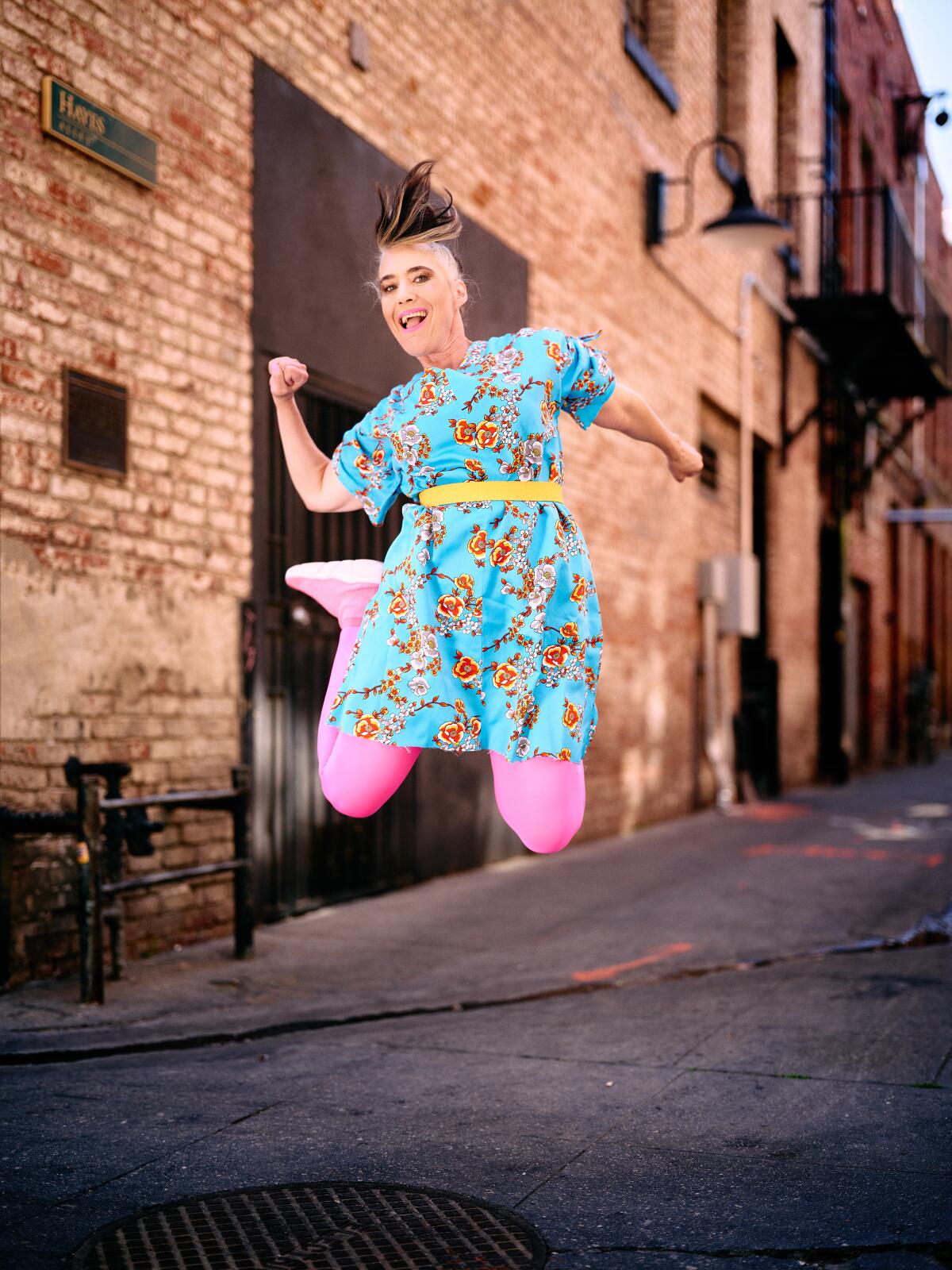 Kathleen Hanna, wearing pink tights and a blue floral dress, leaps into the air.