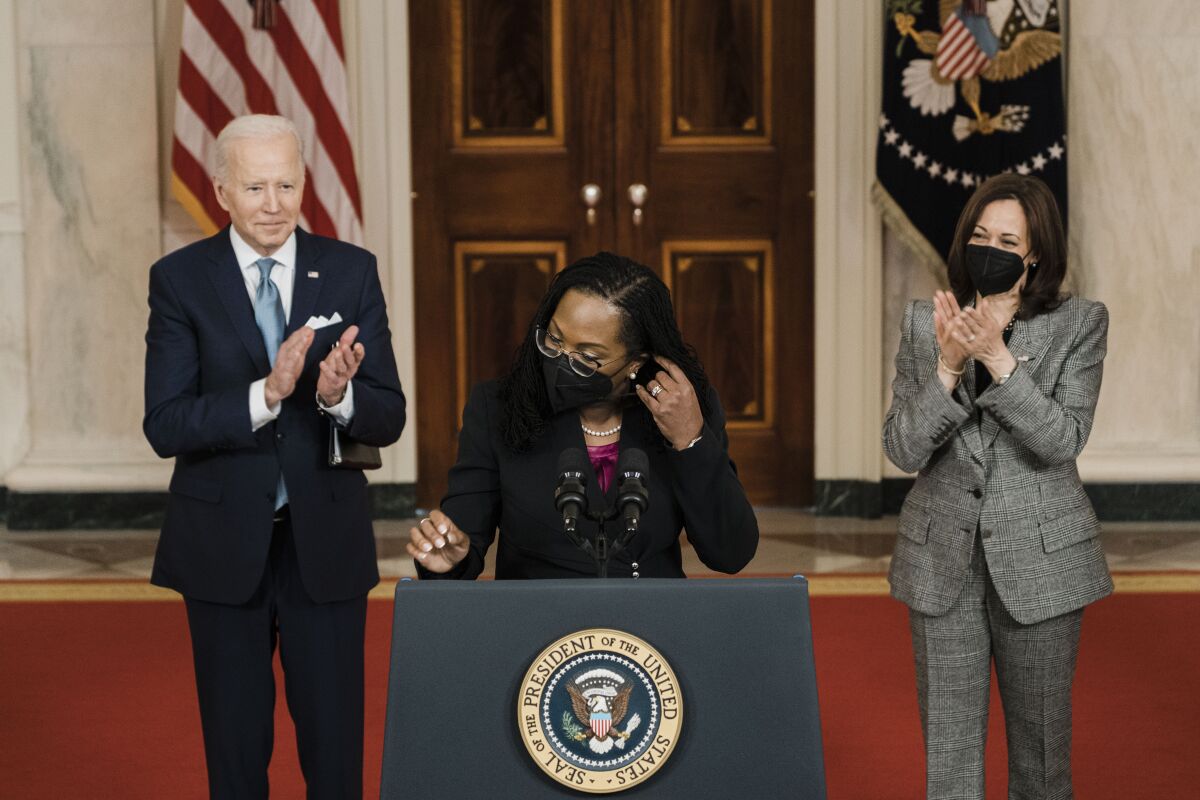 Judge Ketanji Brown Jackson removes her mask at a lectern as President Biden and Vice President Kamala Harris applaud her.